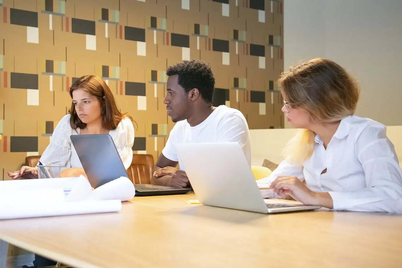 Разнообразная группа дизайнеров совместно работает над проектом: они сидят за столом с ноутбуками и чертежами и разговаривают.